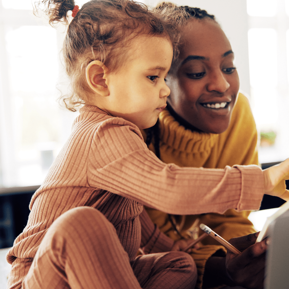 A child in a pink outfit sits next to an adult in a yellow sweater. They are both smiling and focused on a tablet screen in front of them. The background is softly blurred, suggesting they are indoors.