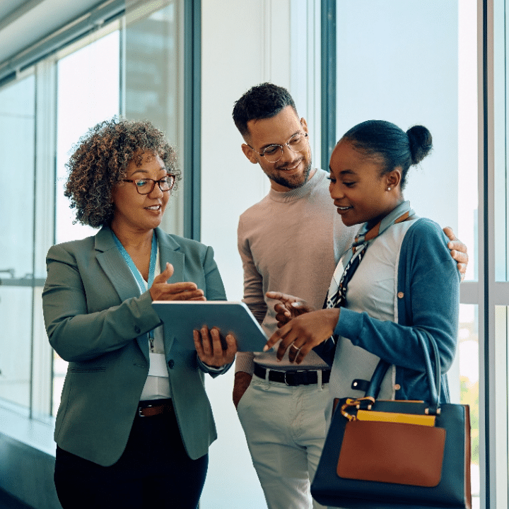 Three colleagues stand by a window, discussing content on a tablet held by one of them. They appear engaged and collaborative, dressed in business-casual attire. Bright daylight streams in, suggesting a modern office setting.
