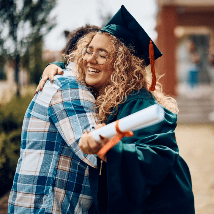A graduate in a cap and gown, holding a diploma, joyfully hugs a person wearing a plaid shirt. They are outside, and the background is slightly blurred. The graduate is smiling, showing excitement and achievement.