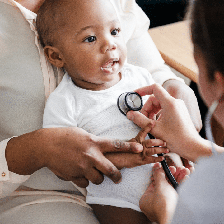 A baby in a white onesie sits on an adult's lap while a medical professional uses a stethoscope to listen to the baby's chest. The baby is looking up with an open mouth, appearing curious and engaged.