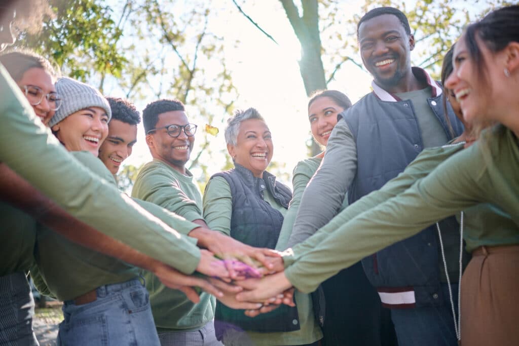 A diverse group of people stands in a circle outdoors, smiling and stacking their hands together in the center. They are surrounded by trees and sunlight filters through the leaves.