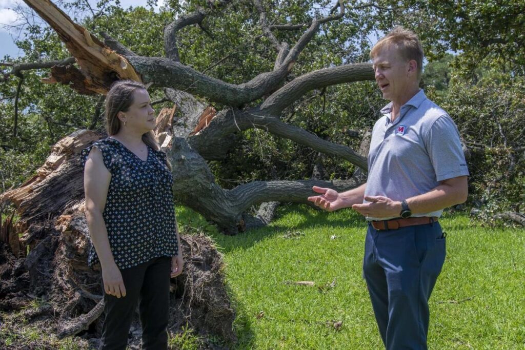 A man and a woman stand on grass near a large fallen tree with exposed roots. The man gestures as he speaks, and the woman listens attentively. They appear to be discussing the aftermath of the tree fall.